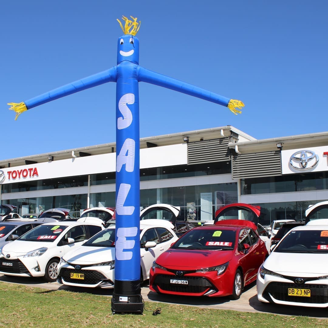 112SAB1 Blue inflatable advertising man waving in front of Toyota car dealership and vehicles.